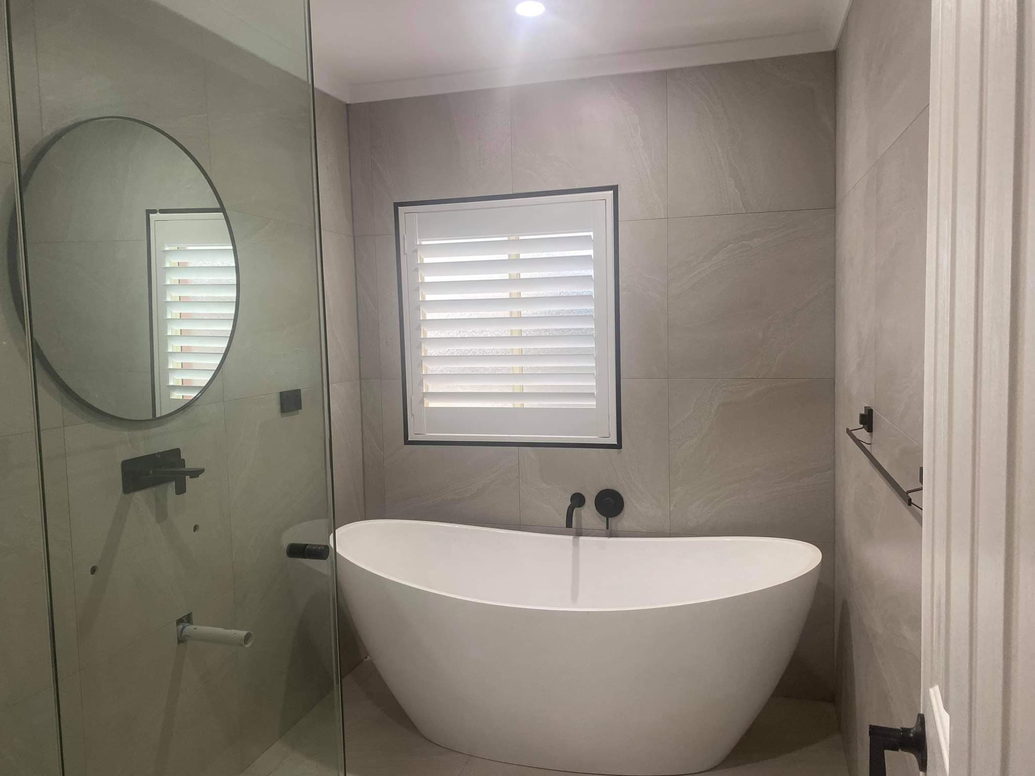 White PVC plantation shutters above a freestanding bath in a grey tiled bathroom